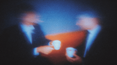 Two figures across a table in a softly blurred meeting scene.